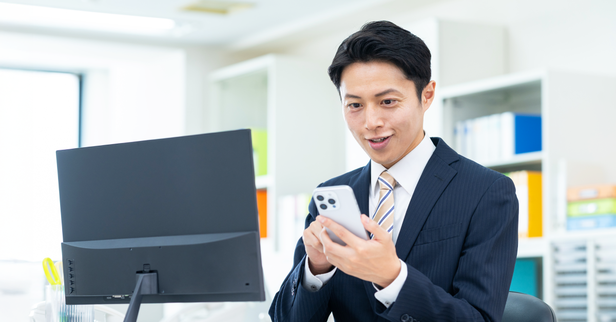 オフィス内でPCに向けてスマホを操作している写真イメージ画像